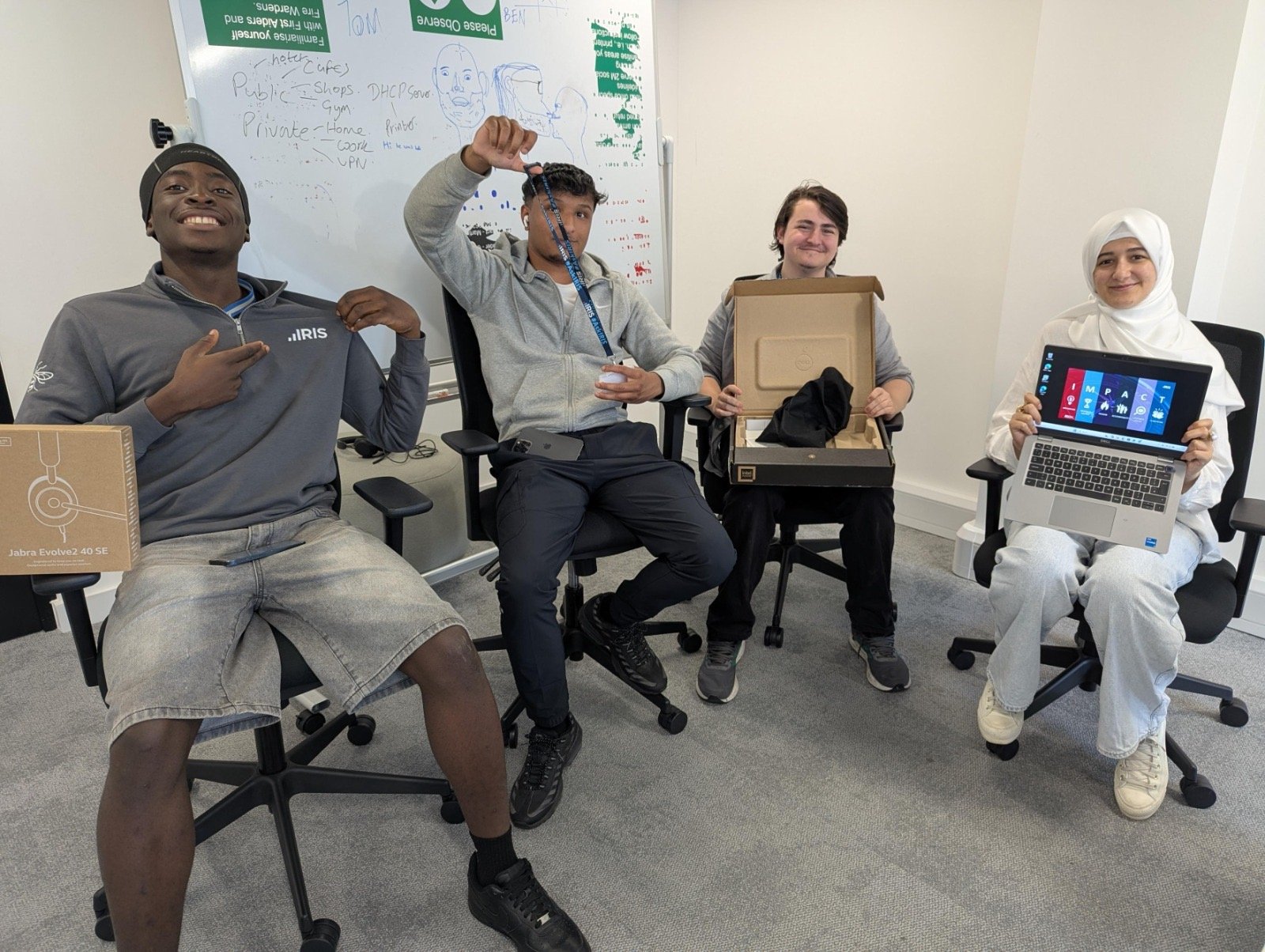 Four IRIS Software Group interns sitting in office chairs, smiling at the camera, with a headset box, an open laptop box and a laptop.